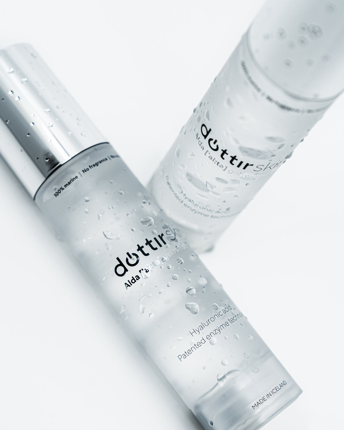 Two clear cosmetic bottles with water droplets, one with a silver cap in the foreground and one in the background, labeled Dóttir Skin, Alda gel moisturiser. Shot on a clean white background. Hyaluronic acid. Patented enzyme technology. 100% marine, no fragrance no nonsense. MADE IN ICELAND. 
